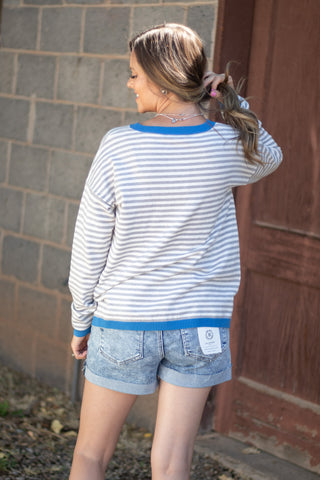 A woman shown from behind wearing a white and blue striped long-sleeve t-shirt and denim shorts, standing outdoors next to a gray brick wall and a brown wooden door.