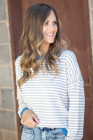 A woman with long wavy brown hair wearing a light blue and white striped long-sleeve shirt and blue jeans, standing in front of a rustic brown wooden door.