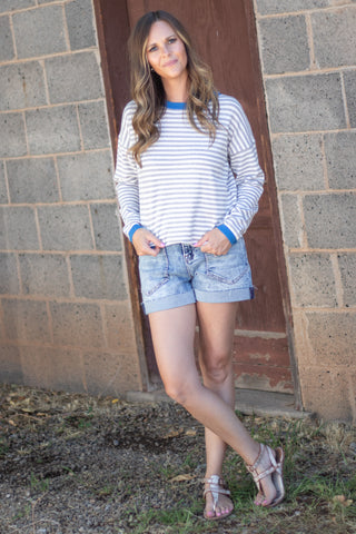 A woman stands outdoors in front of a brick wall and wooden door, wearing a white long-sleeve striped shirt with blue trim, light-wash denim shorts, and beige sandals.