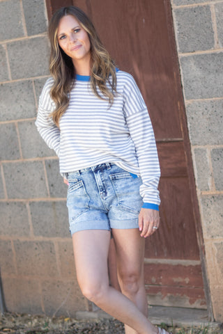 A woman wearing a blue and white striped long-sleeve t-shirt and light-wash denim shorts, posing in front of a gray brick wall and a brown wooden door.