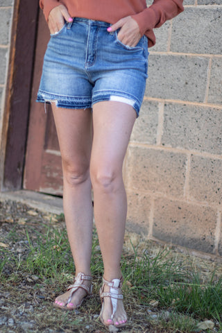 A person wearing light-wash blue denim cutoff shorts with a raw hem, paired with a rust-colored long-sleeve top and beige strappy sandals, standing outdoors against a cinderblock wall.