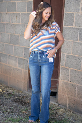 A woman wearing a blue striped short-sleeve t-shirt and blue flare jeans, standing outdoors in front of a gray brick wall.