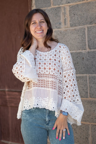 A woman wearing a white eyelet crochet long-sleeve blouse with blue jeans, standing in front of a gray stone wall.