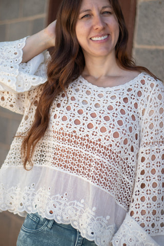 A woman smiling wearing a natural colored crochet and embroidered long sleeve top with acid green jeans. 