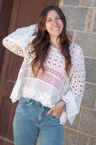 A woman wearing a white crochet lace long-sleeve top with blue jeans, posing outdoors against a stone wall and wooden door.