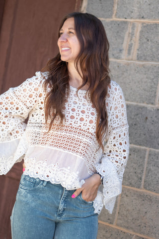 A woman wearing a white eyelet lace long-sleeve blouse and green jeans, standing outdoors against a gray stone wall and brown wooden door.