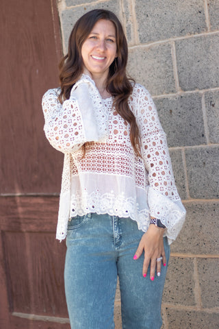 A woman wearing a white long-sleeve lace blouse with a sheer patterned bodice and green jeans, standing in front of a gray stone wall and a brown wooden door.