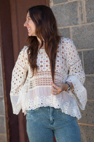 A woman smiling wearing a natural colored crochet and embroidered long sleeve top with acid green jeans. 
