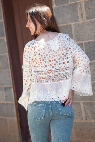 A woman smiling wearing a natural colored crochet and embroidered long sleeve top with acid green jeans. 
