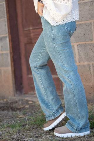 A person wearing light-wash blue flare jeans, a white lace-trim blouse, and brown slip-on shoes, standing outdoors against a brick wall and wooden door.