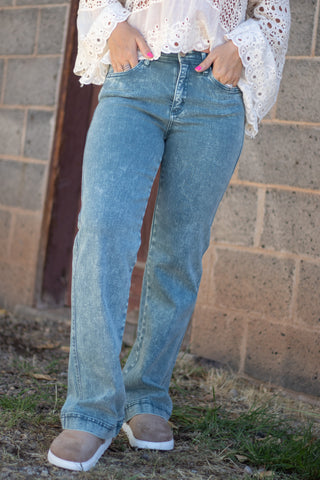 A person wearing light-wash blue straight-leg jeans, a white lace blouse, and tan slip-on shoes, standing outdoors against a brick wall.