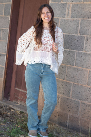 A woman wearing a white crochet long-sleeve top and blue jeans stands outdoors against a gray brick wall.