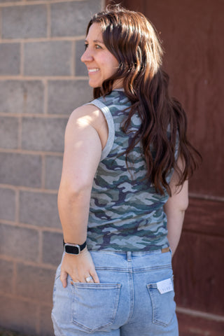 Woman wearing a camouflage tank top and blue jeans standing next to an American flag against a gray wall.