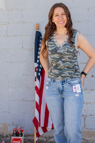 Woman wearing a camouflage tank top and blue jeans standing next to an American flag against a gray wall.