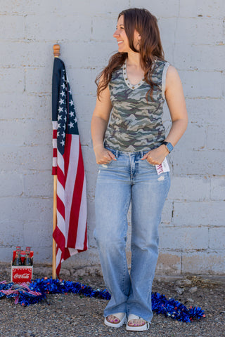 Woman wearing a camouflage tank top and blue jeans standing next to an American flag against a gray wall.