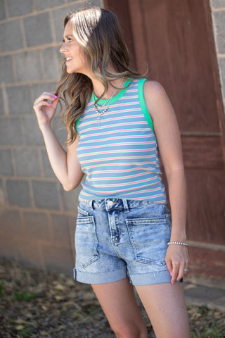 A woman wearing a light blue and white striped tank top with bright green trim and light-wash denim shorts, standing outdoors against a brick wall.