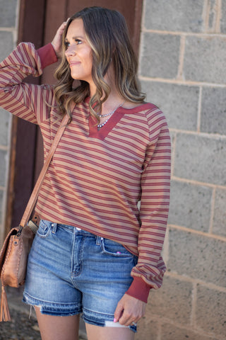 A woman wearing a rust and red striped long-sleeve top with a V-neck, paired with denim shorts and a tan crossbody bag, standing against a gray brick wall.