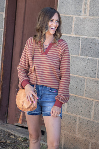 A woman wearing a rust and beige striped long-sleeve V-neck sweater, denim shorts, and carrying a tan leather crossbody bag, standing against a gray brick wall.