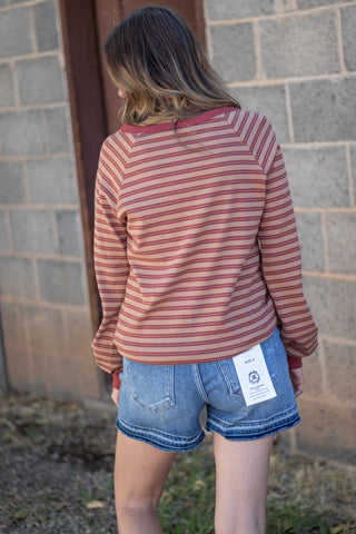 A woman viewed from behind wearing a dusty rose and red striped long-sleeve raglan shirt and denim shorts, standing outdoors against a gray brick wall.