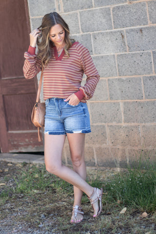 A woman stands outdoors in front of a gray cinderblock wall, wearing a rust and beige striped long-sleeve knit top, blue denim shorts, and strappy flat sandals. She is carrying a tan crossbody bag.