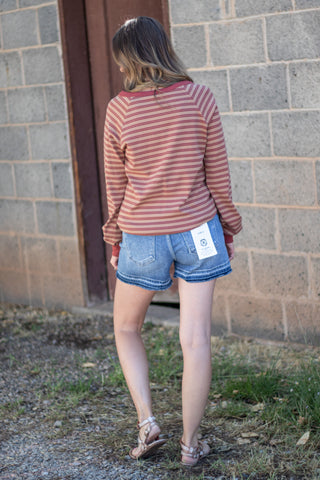 A woman viewed from behind wearing a rust-orange and pink striped long-sleeve shirt, denim shorts, and sandals, standing outdoors against a gray brick wall.