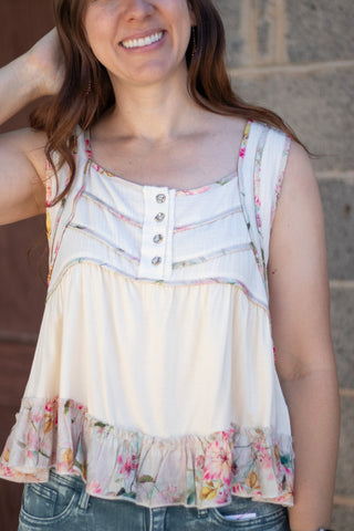 A woman wearing a sleeveless cream-colored top with a floral ruffle hem and decorative buttons on the chest, paired with green jeans, standing against a stone wall.