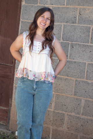 A woman wearing a sleeveless cream-colored top with a floral ruffle hem and decorative buttons on the chest, paired with green jeans, standing against a stone wall.