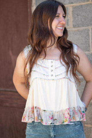 A woman wearing a sleeveless cream-colored top with a floral ruffle hem and decorative buttons on the chest, paired with green jeans, standing against a stone wall.