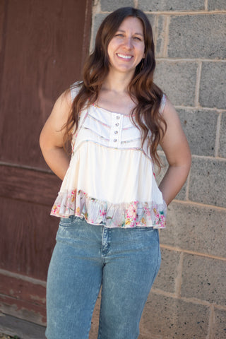 A woman wearing a sleeveless cream-colored top with a floral ruffle hem and decorative buttons on the chest, paired with green jeans, standing against a stone wall.