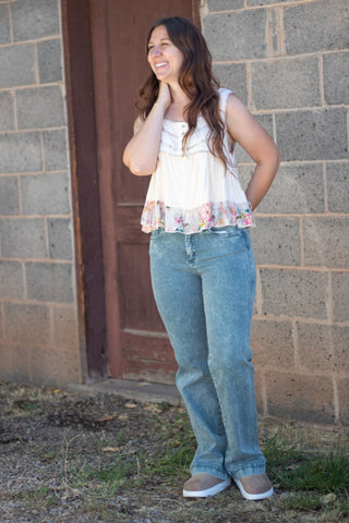 A woman wearing a sleeveless cream-colored top with a floral ruffle hem and decorative buttons on the chest, paired with green jeans, standing against a stone wall.
