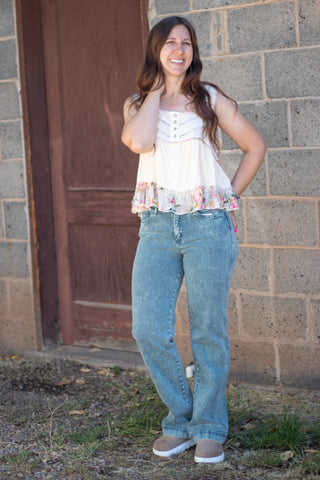 A woman wearing a sleeveless cream-colored top with a floral ruffle hem and decorative buttons on the chest, paired with green jeans, standing against a stone wall.