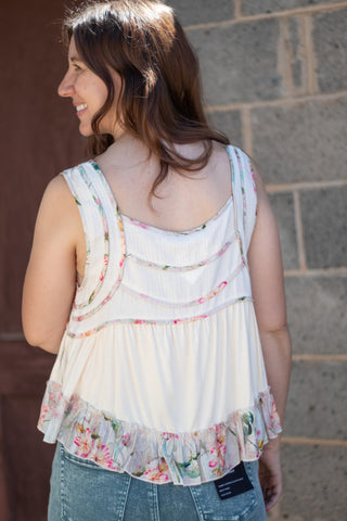 A woman wearing a sleeveless cream-colored top with a floral ruffle hem and decorative buttons on the chest, paired with green jeans, standing against a stone wall.
