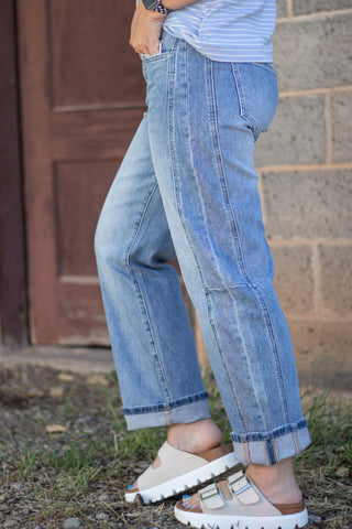 A person wearing light blue wide-leg denim jeans with cuffed hems, paired with beige slide sandals, standing outdoors in front of a rustic brown door and stone wall.