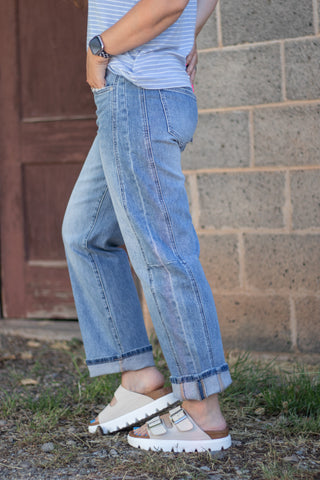 A person wearing light blue wide-leg denim jeans with rolled cuffs, paired with a striped top and beige slide sandals, standing outdoors against a brick wall.
