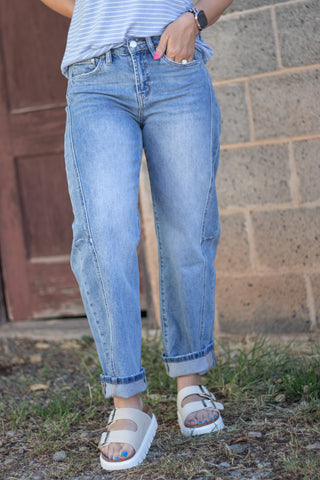 A person wearing light-wash blue denim jeans with rolled cuffs, paired with a striped top and white slide sandals, standing outdoors against a brick wall and wooden door.