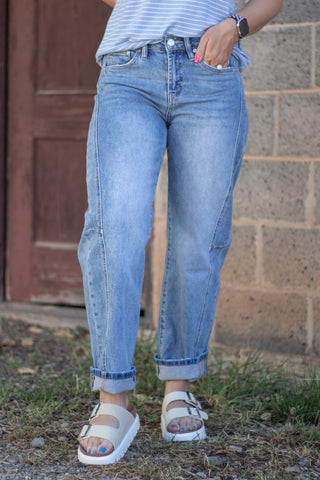 A person wearing light-wash blue jeans with rolled cuffs, paired with white slide sandals and a striped top, standing outdoors in front of a brick wall and wooden door.
