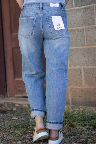 Rear view of a person wearing light blue denim jeans with rolled cuffs and white slip-on shoes, standing outdoors against a brick wall and wooden door.