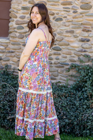 Woman wearing a colorful floral dress standing outdoors with a stone wall and greenery in the background.