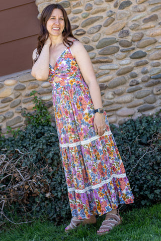 Woman wearing a colorful floral dress standing outdoors with a stone wall and greenery in the background.