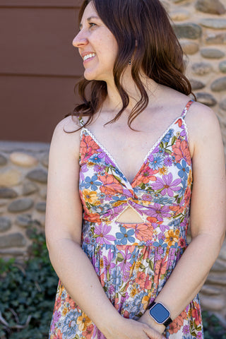 Woman wearing a colorful floral dress standing outdoors with a stone wall and greenery in the background.