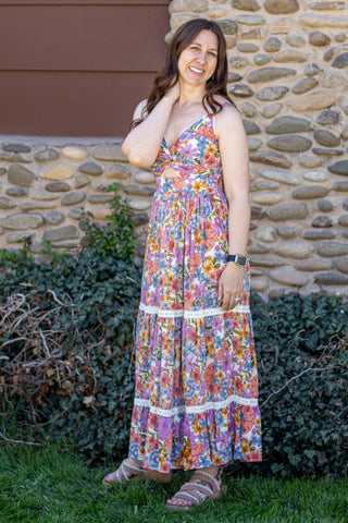Woman wearing a colorful floral dress standing outdoors with a stone wall and greenery in the background.