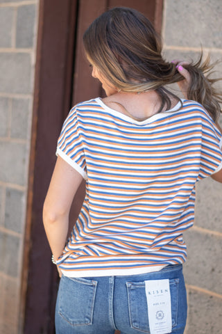 Woman wearing a blue, copper and white striped short sleeve top with a pair of dark wash denim. 