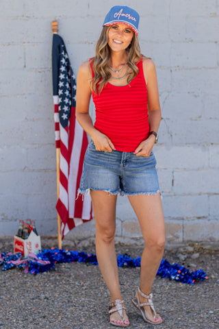 Woman wearing a red tank top, denim shorts, and a blue cap with 'America' text in front of an American flag.