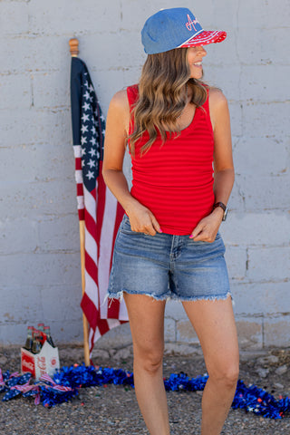 Woman wearing a red tank top, denim shorts, and a blue cap with 'America' text in front of an American flag.