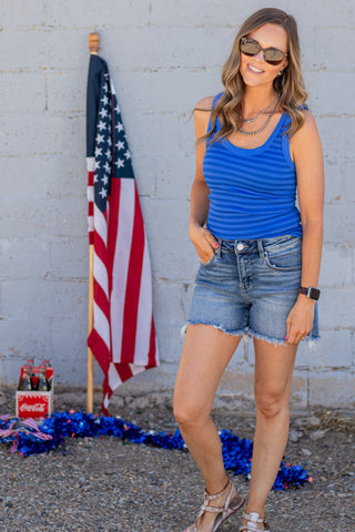 Woman wearing a blue striped tank top and denim shorts standing in front of an American flag.