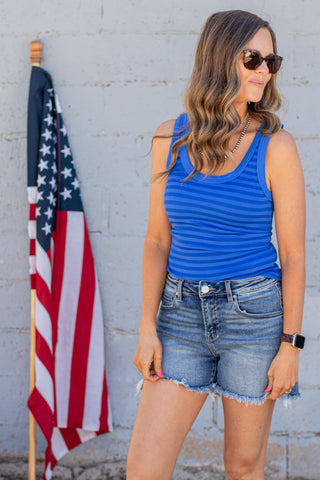 Woman wearing a blue striped tank top and denim shorts standing in front of an American flag.