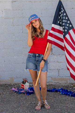 Woman wearing a red tank top, denim shorts, and a blue cap with 'America' text in front of an American flag.