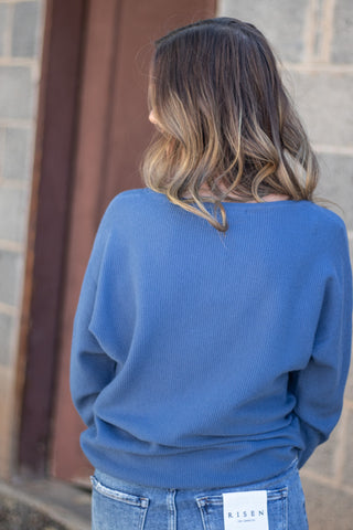 A woman viewed from behind wearing a blue ribbed knit sweater and blue jeans, standing outdoors against a stone wall and wooden door.