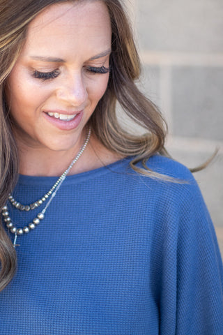 A close-up lifestyle shot of a woman wearing a textured blue knit top and a double-strand silver beaded necklace, looking down with a soft smile.