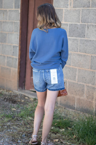 A woman viewed from behind wearing a blue long-sleeve sweatshirt and light-wash denim shorts, standing outdoors against a gray brick wall.